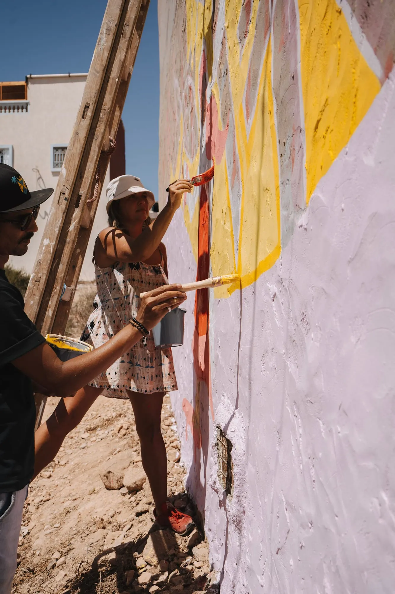 Two people painting a large outdoor wall in yellow, pink and orange tones under the sun