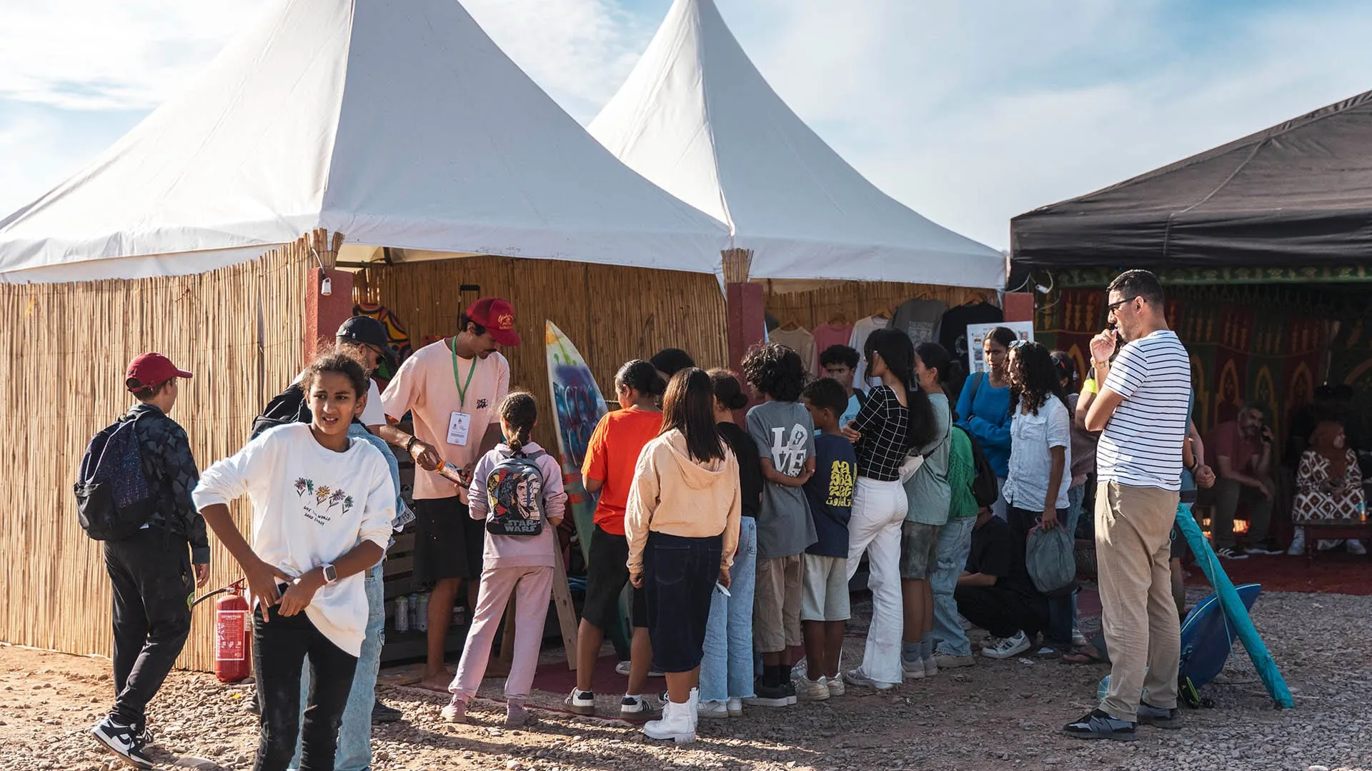 Children lining up at a festival tent for a street-art workshop, a painted surfboard leaning against the stand