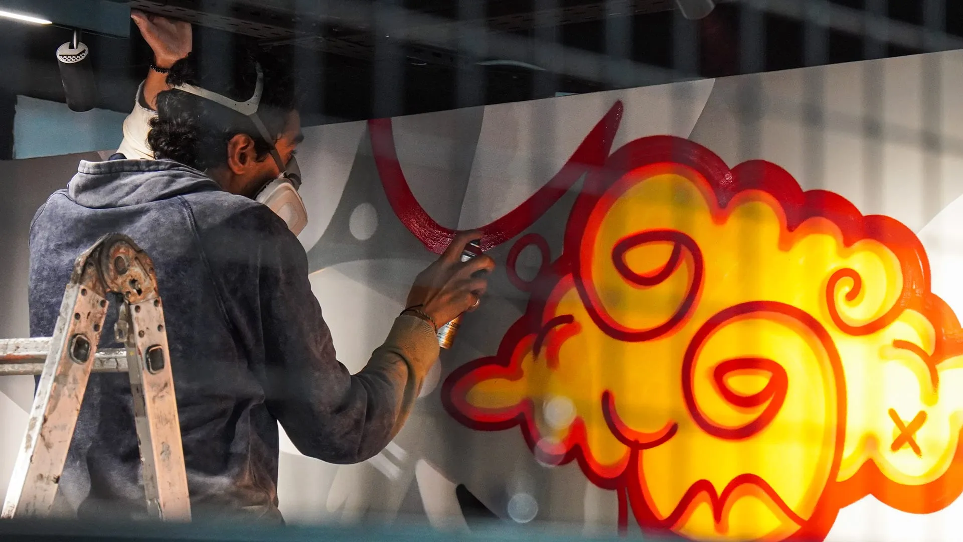 Zaamom wearing a respirator mask, spray painting a yellow and red cloud character on a wall at night