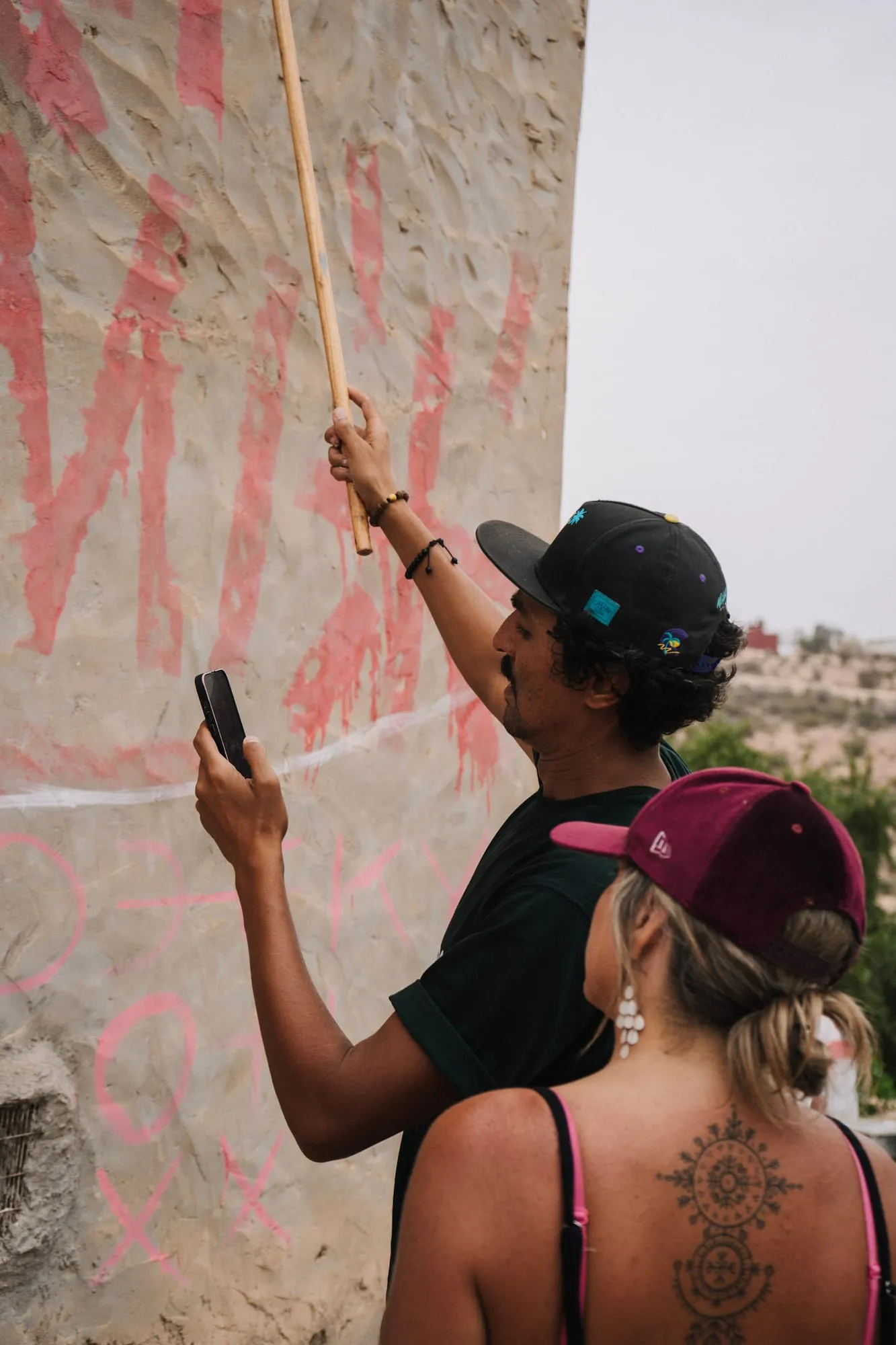 Zaamom tracing the first large outlines of a mural on an outdoor wall, phone in hand